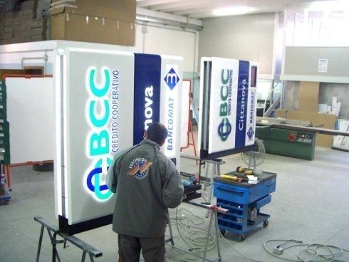 Un uomo lavora alle insegne luminose di una banca in un'officina. Sono visibili i loghi