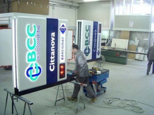 Due uomini lavorano alle insegne luminose di una banca in un'officina; le insegne mostrano i loghi