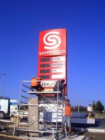 Operai che installano un cartello per una stazione di servizio Sanrocco. Cartello rosso e bianco, cielo azzurro.