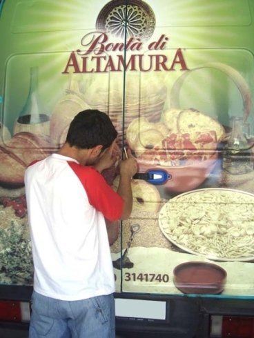 Uomo ispeziona la portiera di un furgone con una torcia; sul furgone c'è la pubblicità