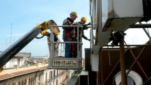 Due operai edili, in un elevatore a cestello, con caschi e imbracature, lavorano all'esterno di un edificio.