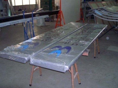 Due grandi pannelli argentati con loghi blu, avvolti nella plastica, appoggiati su cavalletti in un'officina.