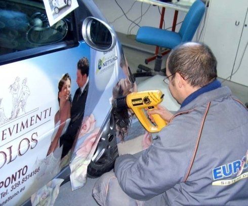 Un uomo applica una pellicola protettiva a un'auto con una pistola termica. La pellicola è a tema matrimonio con coppie.