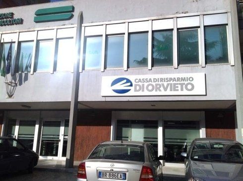 Exterior of Cassa di Risparmio di Orvieto bank with cars parked in front. Gray building with logo above entrance.