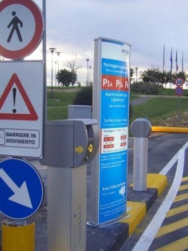 Area di parcheggio con segnaletica, tra cui un cartello blu, un cartello pedonale e un parchimetro.