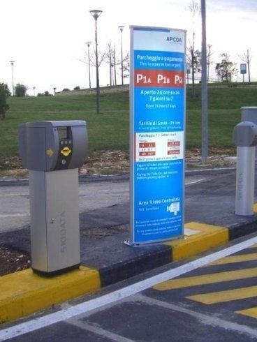 Cartello di parcheggio con cassa automatica. Il cartello è blu e bianco, la cassa automatica è grigia. Marciapiede giallo, asfalto.