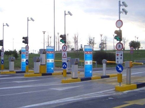 Ingresso al parcheggio con più stazioni di pagamento automatiche, barriere e semafori; segnaletica con pannelli blu e bianchi.