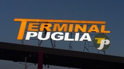 Cartello per il Terminal Puglia, con lettere gialle e bianche su sfondo blu cielo. Include un logo verde e giallo.