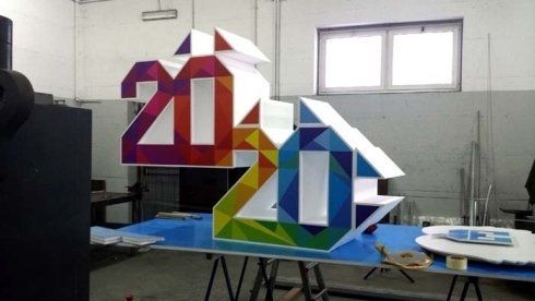 Grande e colorato cartello geometrico con la scritta 2020; sfumature dal rosso al viola e dal giallo al blu; in un laboratorio, vicino alla finestra.