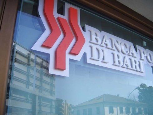 Logo della Banca Popolare di Bari, rosso e bianco su una vetrata, con riflessi di edifici.