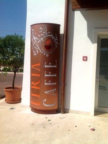 Insegna marrone per Liria Caffè con il disegno di una tazzina da caffè, accanto a un muro bianco e un albero in vaso.