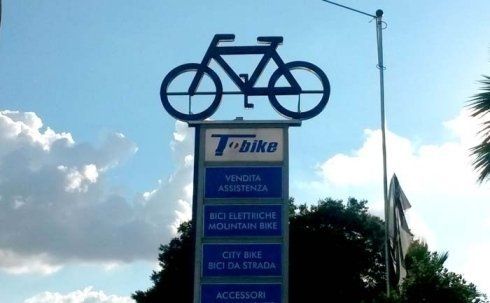 Cartello per T-bike con la sagoma di una bicicletta in alto. Sotto, l'elenco dei servizi in italiano, sullo sfondo di un cielo azzurro.