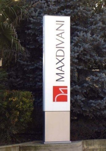 Cartello per Maxdivani con sfondo bianco, testo nero e logo rosso, esposto all'esterno tra gli alberi.