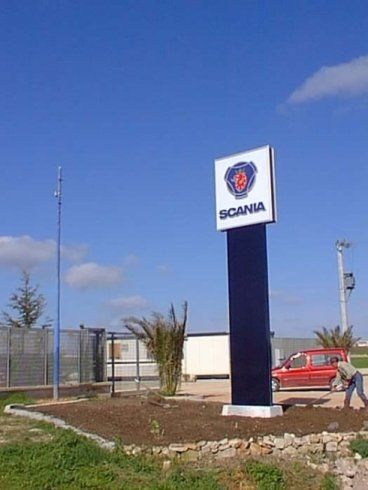 Cartello Scania presso una concessionaria, con un operaio impegnato in giardinaggio di fronte e cielo azzurro sullo sfondo.