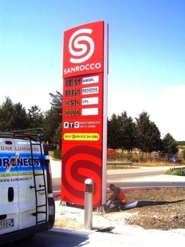 Un cartello rosso di una stazione di servizio Sanrocco che mostra i prezzi. Un operaio con un gilet arancione è vicino alla base.