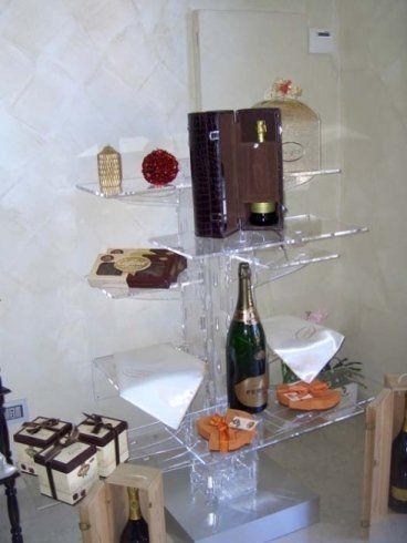 Mensola espositiva in acrilico trasparente con varie scatole regalo, bottiglie di champagne e altri articoli decorativi in un ambiente di vendita al dettaglio.