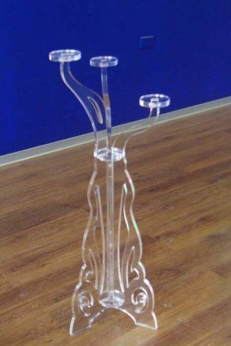 Portacandele in acrilico trasparente con tre bracci, appoggiato su un pavimento in legno contro una parete blu.