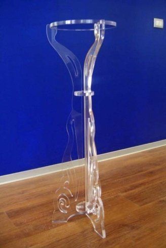 Piedistallo in acrilico trasparente appoggiato su una parete blu e un pavimento in legno.