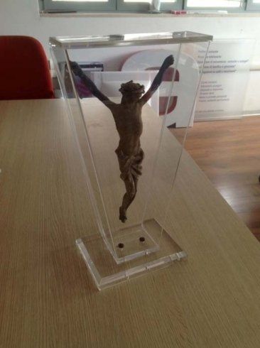 Statua in legno di Gesù in croce all'interno di una teca in acrilico trasparente, appoggiata su un tavolo di legno.