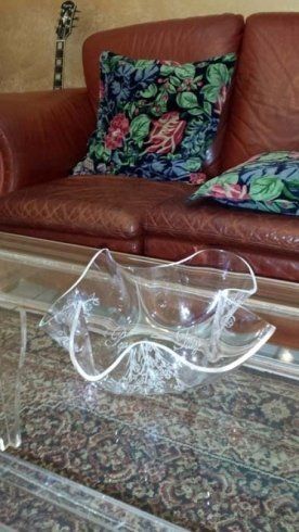Ciotola di vetro trasparente e ondulata su un tavolo di vetro di fronte a un divano in pelle rossa con cuscini floreali.