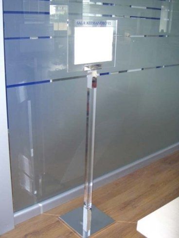 Portacartello con insegna bianca vuota davanti a una parete di vetro. Palo cromato su base quadrata, per interni.