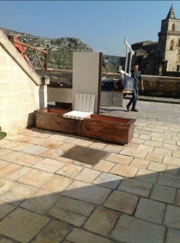 Uomo che cammina accanto a una panchina di legno su un patio in pietra, con un edificio e montagne sullo sfondo.