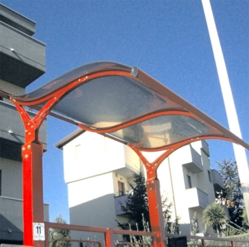 Pensilina con struttura rossa e tetto trasparente, di fronte a condomini, in una giornata di sole.