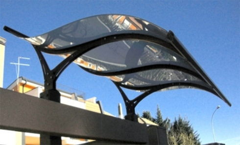 Pensilina metallica sopra un cancello, con tetto trasparente ondulato e struttura portante nera. Cielo azzurro sullo sfondo.