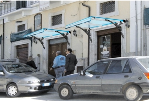 Esterno di un edificio con tende da sole blu, due uomini che entrano. Auto parcheggiate di fronte.