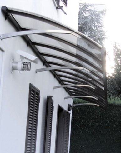 Tenda curva e trasparente sopra la porta e le finestre di un edificio bianco, sostenuta da staffe metalliche.