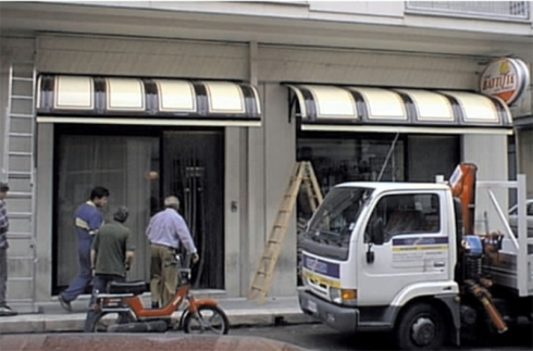 Esterno di un negozio con tende da sole, su cui lavorano diverse persone. Un camion con una gru è parcheggiato nelle vicinanze.