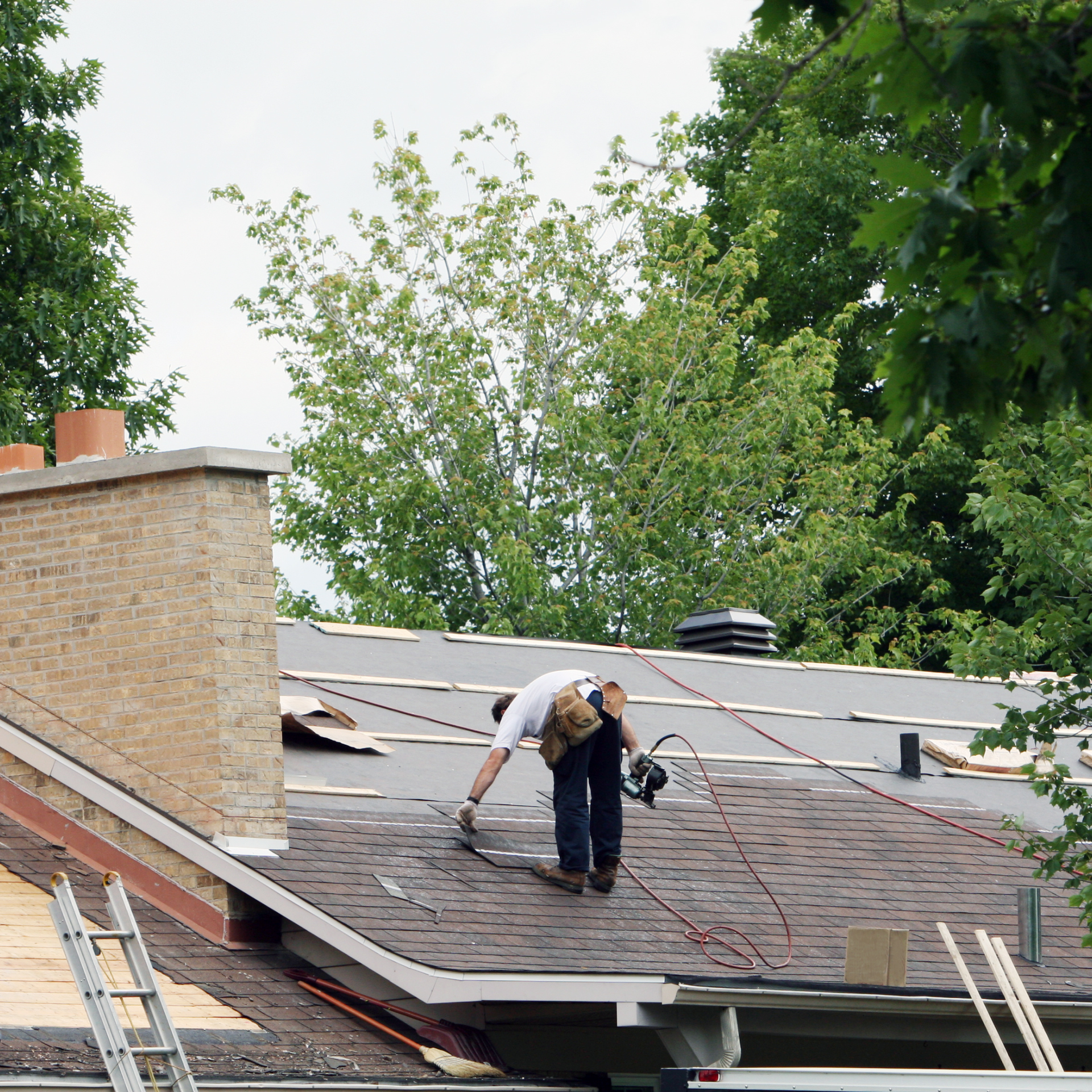Roofer on a residential roof, installing shingles. Brick chimney, ladder, trees in the background.