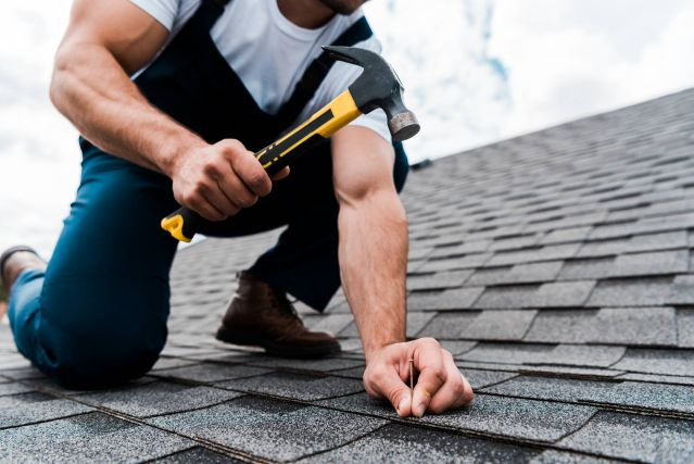 Roofer on a shingled roof, hammering a nail.
