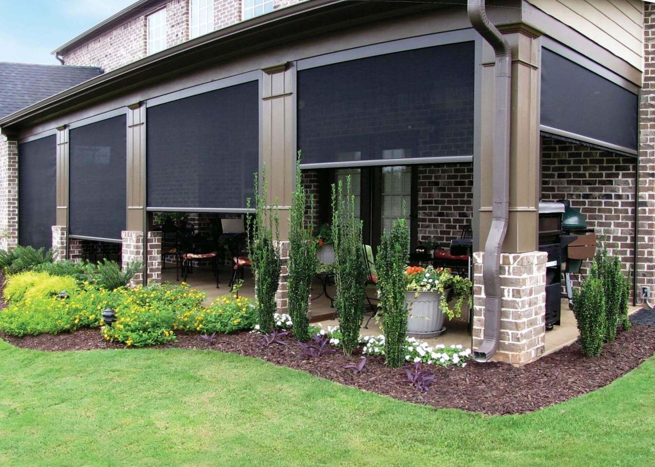 Patio with retractable black sun shades, brick columns, and landscaping. Green grass, shrubs, and flowers border the patio.