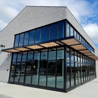 Modern building with large glass windows and a wooden overhang. The exterior has gray siding and a bright sky is reflected in the windows.