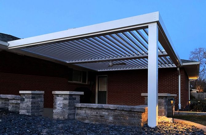 A modern white aluminum pergola covers a brick patio. The pergola has adjustable louvers, and is attached to a red brick house.