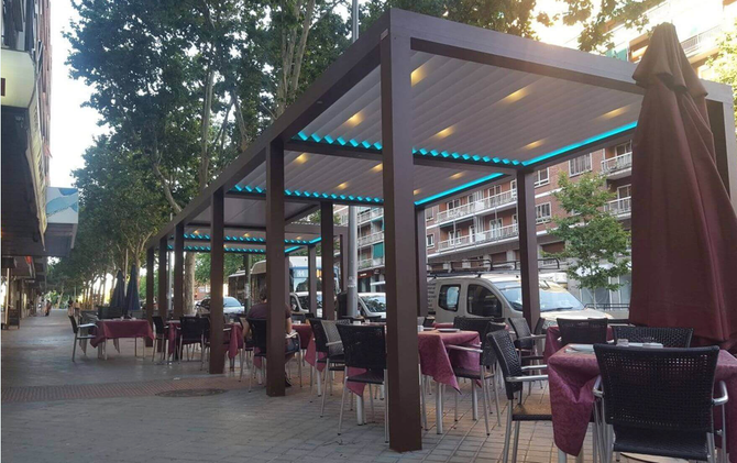 Outdoor restaurant seating with brown pergolas, tables, and chairs along a city sidewalk. Tables have red tablecloths.