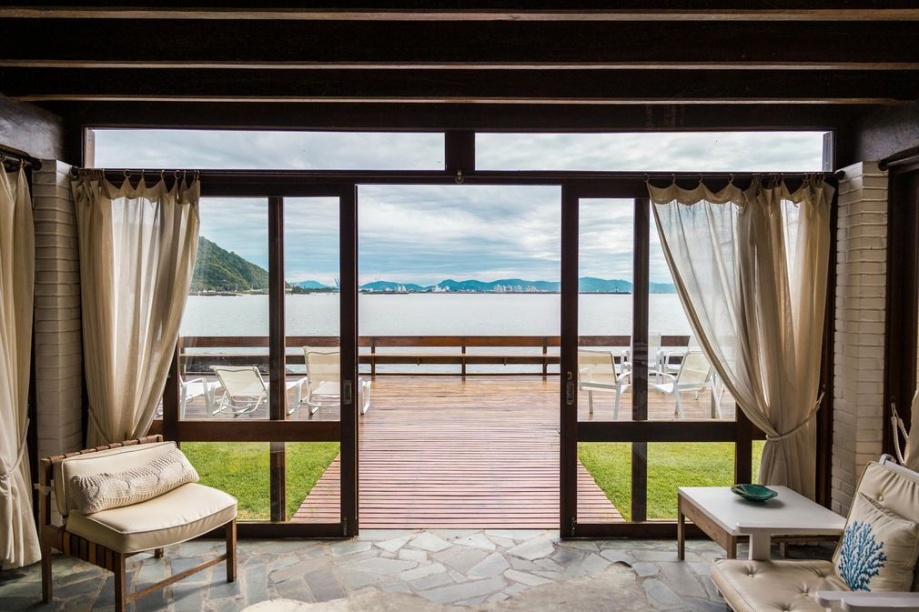 Interior view of a room with open doors leading to a waterfront deck; sheer curtains frame the view of the water and distant buildings.