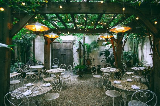 An outdoor restaurant patio with metal tables and chairs under a wooden pergola covered in vines and string lights.