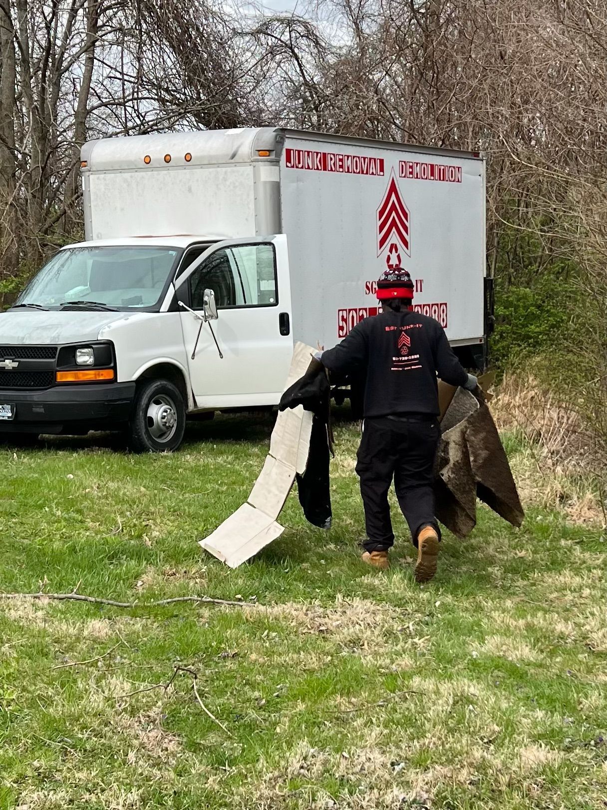 Man carrying items toward a white moving truck parked on grass; truck has logo of an arrow design.