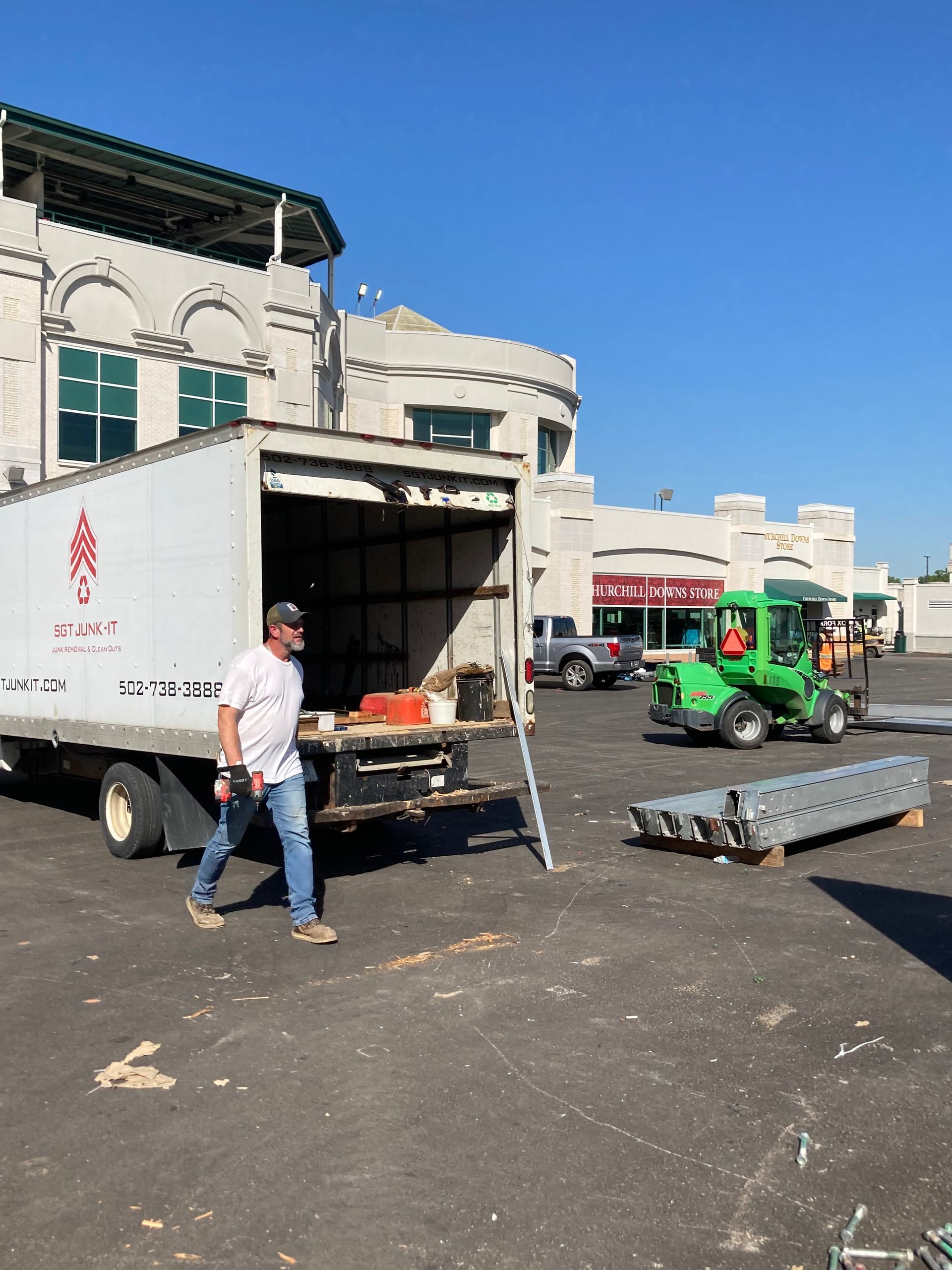 Man exits a box truck; equipment and a green forklift are nearby; brick building in background.