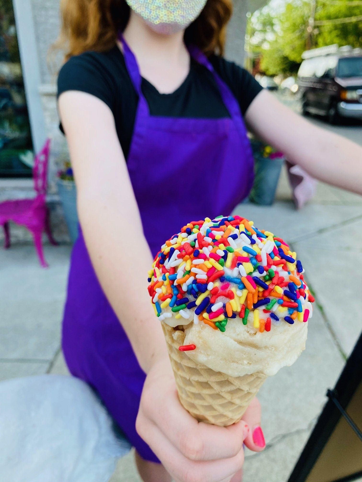 A girl in a purple apron is holding an ice cream cone with sprinkles on top