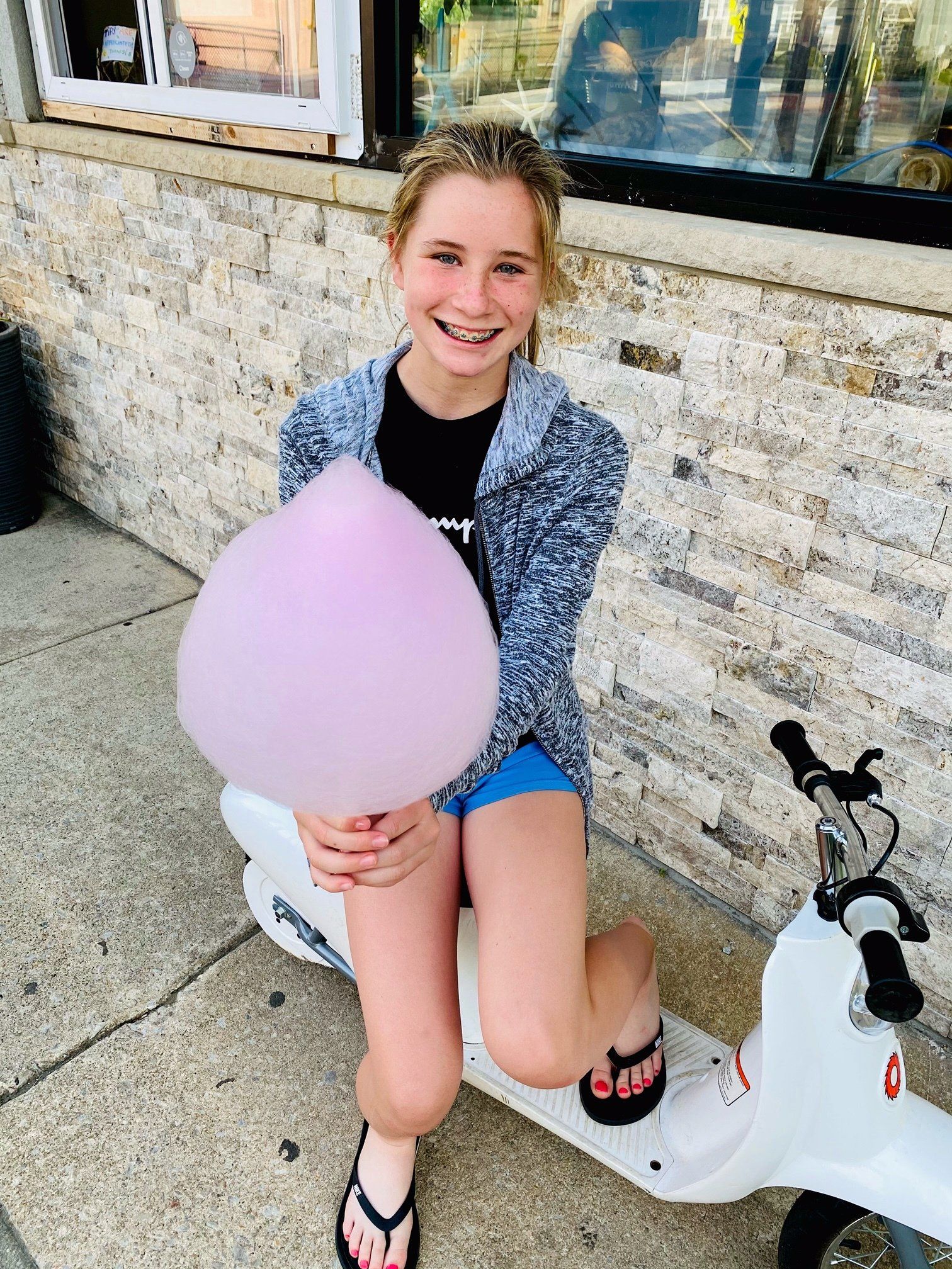 A young girl is sitting on a scooter holding cotton candy.