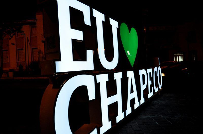 Letreiro luminoso: "EU 💚 CHAPECO" em letras brancas com um coração verde, sobre um fundo escuro.
