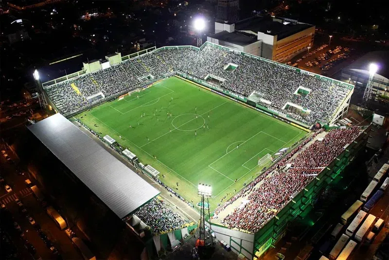 Vista aérea de um estádio de futebol iluminado à noite, lotado de espectadores, campo verde e arquibancadas brancas e verdes.