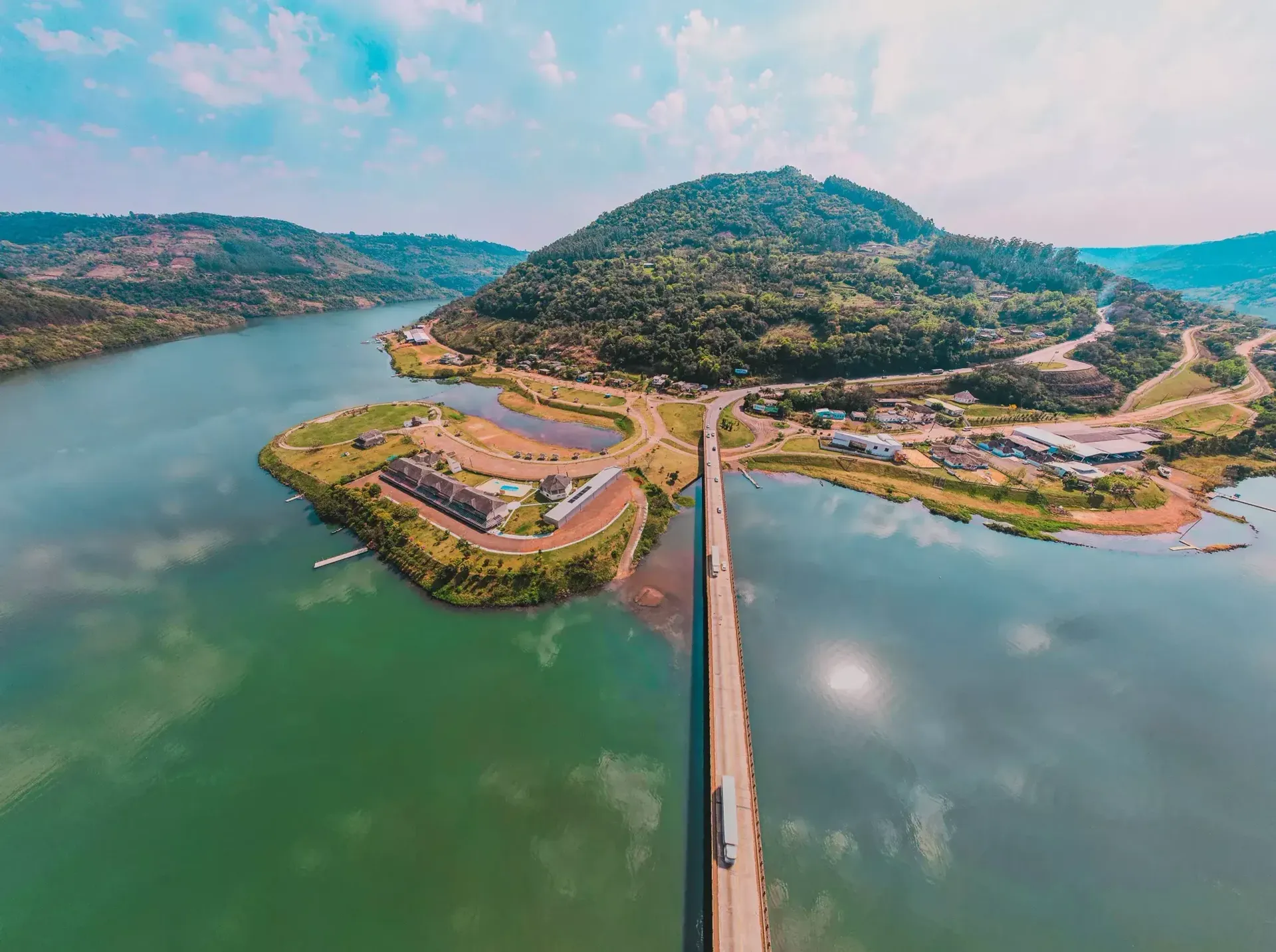 Ponte sobre um lago verde com uma ilha arborizada e colinas circundantes sob um céu azul.