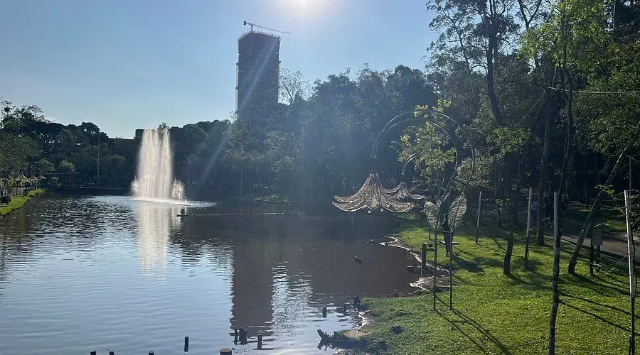 Cena de parque com fonte, lago, árvores e prédio alto sob o sol brilhante.
