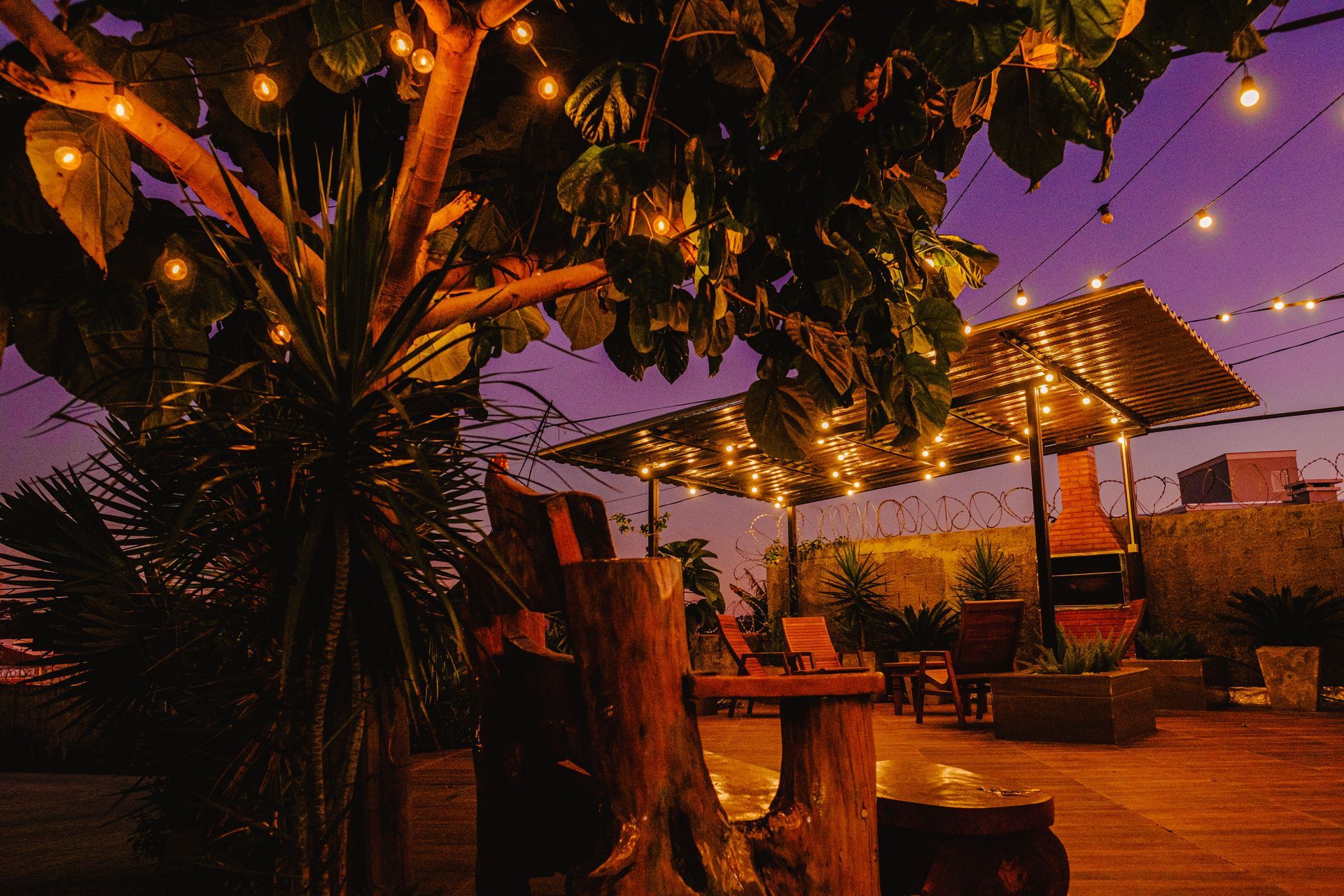 Terraço na cobertura ao entardecer com luzes de corda, árvores, assentos e uma churrasqueira sob um céu roxo.