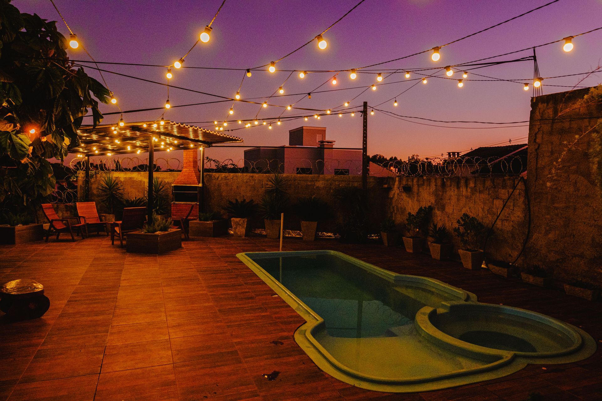 Terraço na cobertura ao entardecer com luzes de corda, pequena piscina e paisagem urbana ao fundo.