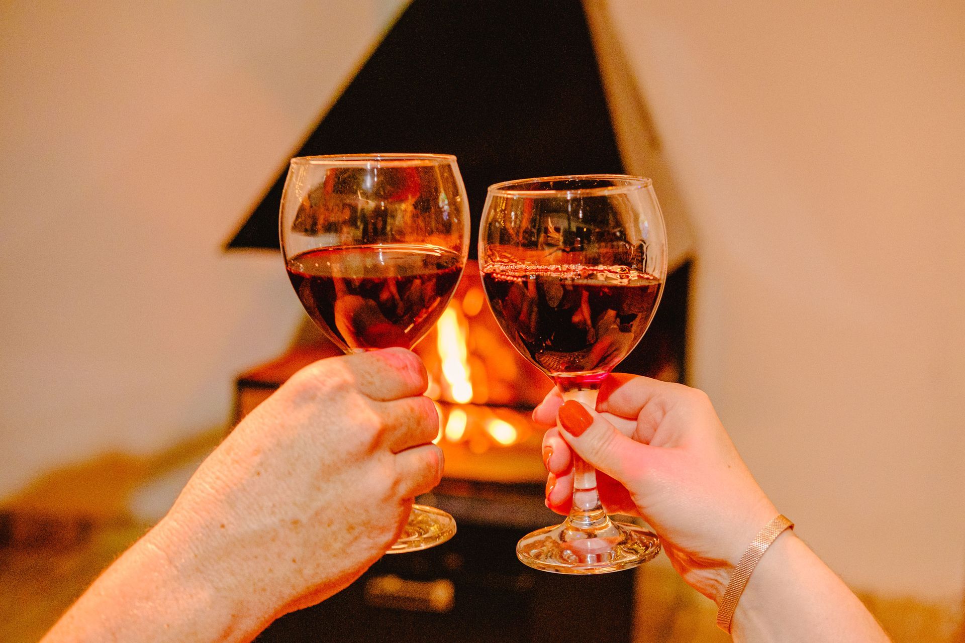 Duas mãos segurando taças de vinho, brindando em frente a uma lareira acesa.